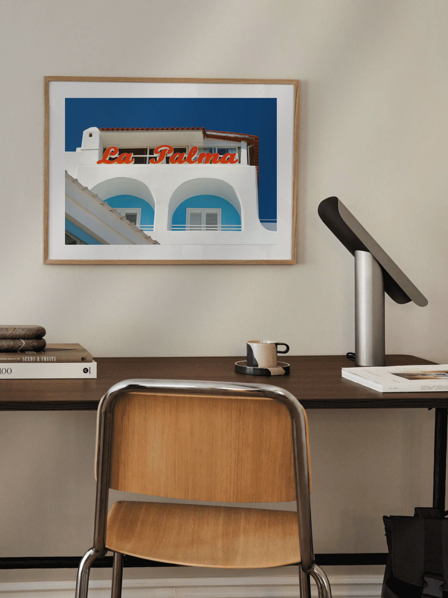 Framed artwork of  Blue and white hotel facade with red sign in daylight above a desk with a chair in a home office setting